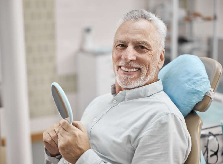Senior man sitting in dental chair holding a hand mirror and smiling. Looking toward camera.