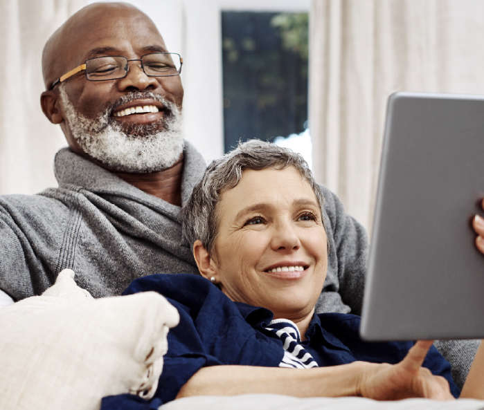 Male female couple sitting on couch smiling. Female leaning against male while holding a tablet.