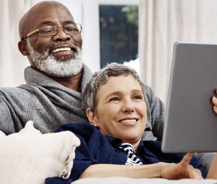 Senior couple sitting on a couch smiling. Woman resting against man while holding a tablet.