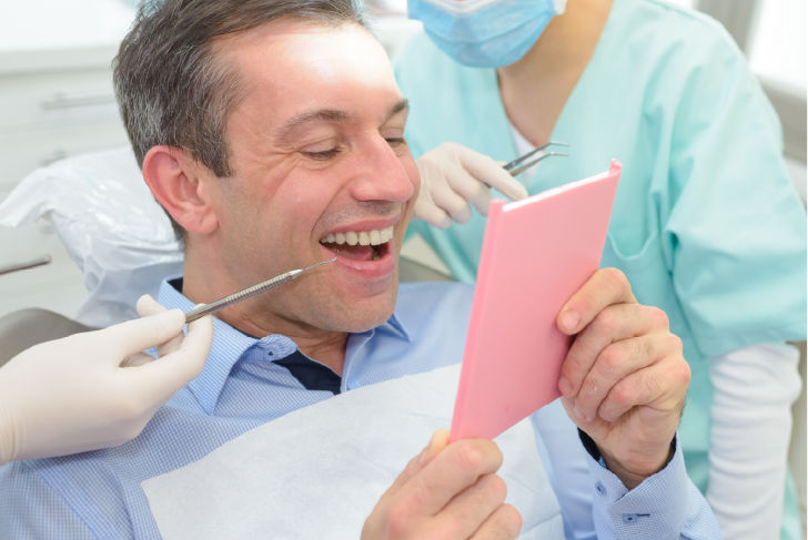 Male patient in dental chair looking in hand mirror at teeth. Periodontist in background.