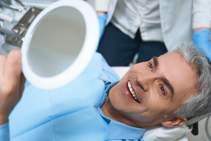 Joyful male lying in chair looking into mirror while delighted with dentist implant