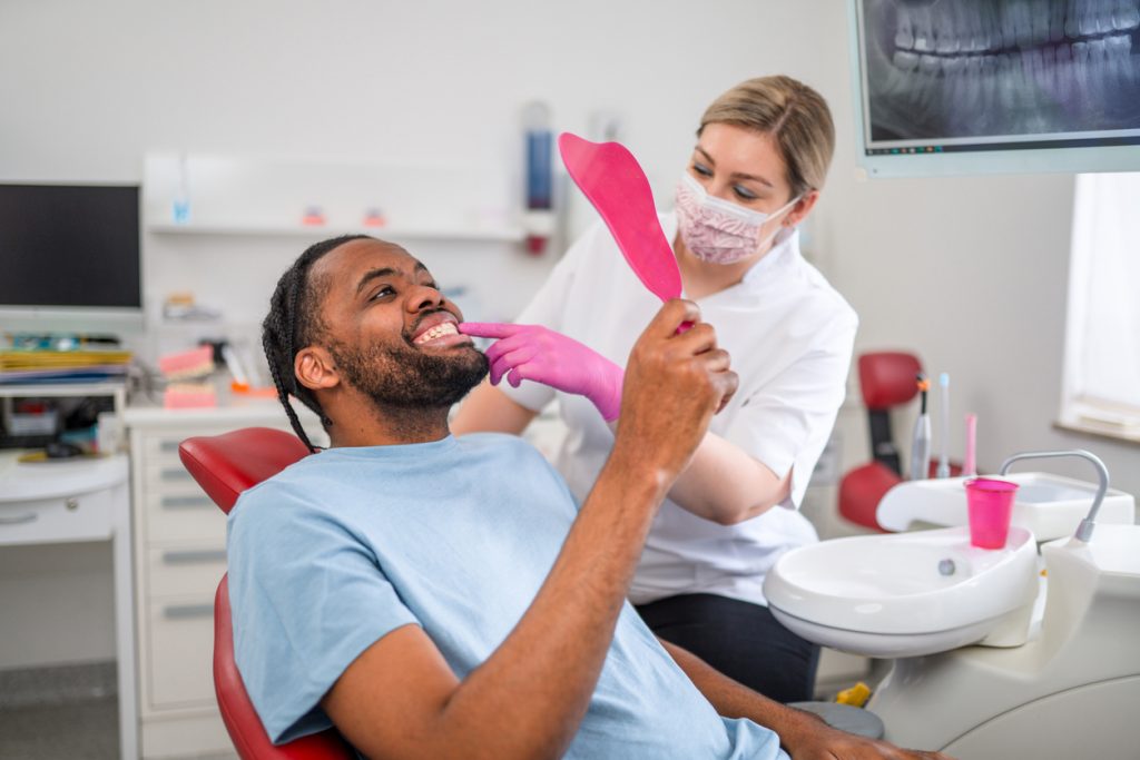 Caucasian female dentist treating tooth of an African-American male patient at dental clinic office.