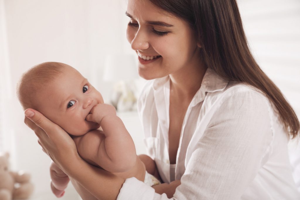 Mother smiling while holding her baby, baby looking at camera with hand in mouth.