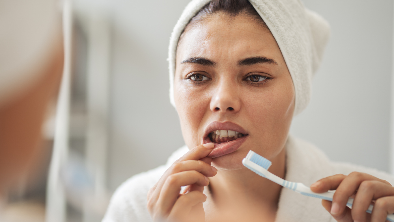 woman with bleeding gums while brushing her teeth looks in the mirror with concern