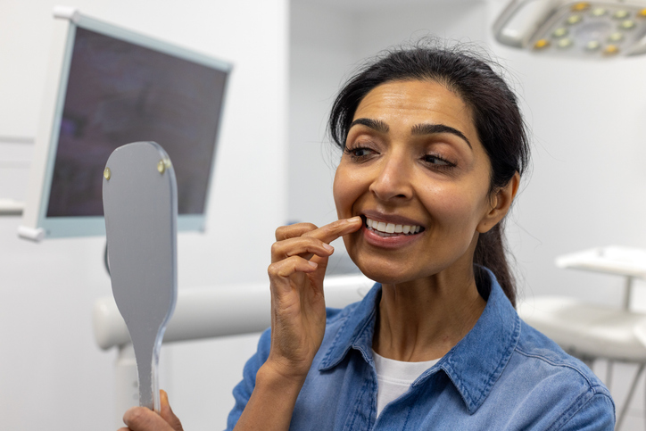 A close-up of an Asian woman holding up a mirror to see her new teeth.